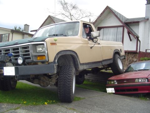 1985 Ford F-150 - A tan Ford pickup truck with a green hood is parked on a driveway, partially blocking a red sedan.