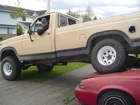 1985 Ford F-150 - A tan pickup truck is positioned on top of a red car, with a person in the driver's seat.