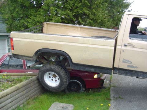 1985 Ford F-150 - A Ford F-150 truck is precariously parked on top of a red car, with its large tires resting on the car's roof.