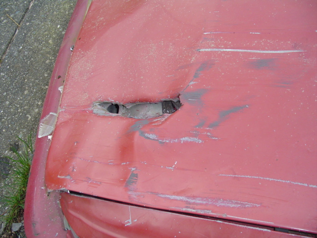 Red vehicle hood with a large hole and dent, showing scraped paint and a broken headlight