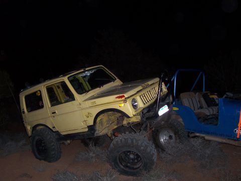 1991 Ford Bronco - Two off-road vehicles are positioned closely together on uneven terrain at night, with one appearing to have sustained front-end damage.