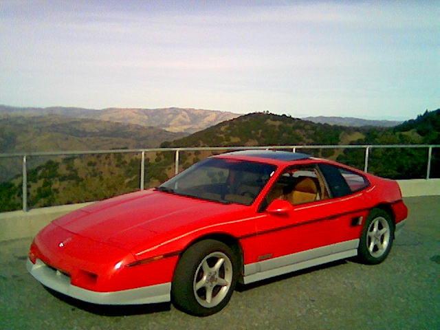 1986 Pontiac Fiero, red, with grey lower body trim, parked with a mountain view
