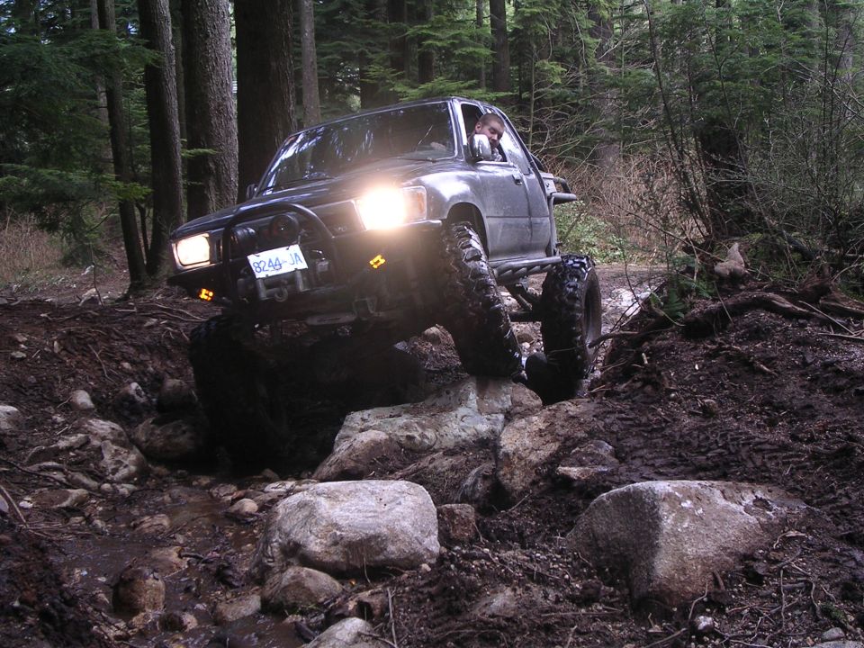 Lifted grey pickup on mud tires crawling over boulders in a forest trail, headlights on