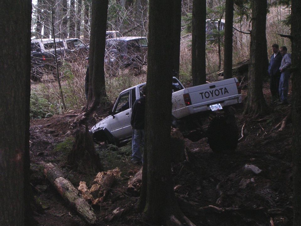 White Toyota pickup truck tilted on a muddy, wooded trail with two people standing nearby