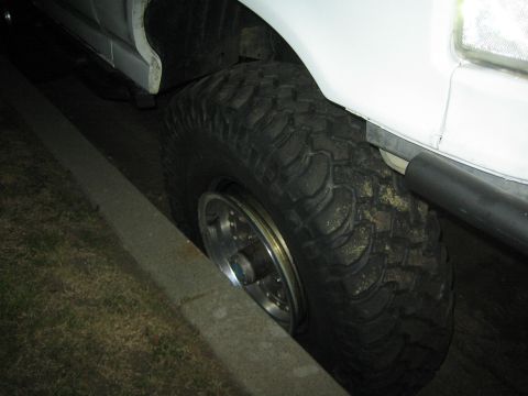 1990 Ford Bronco - Night close-up of a lifted white truck's oversized mud-terrain tire mounted on a polished chrome wheel beside a curb.