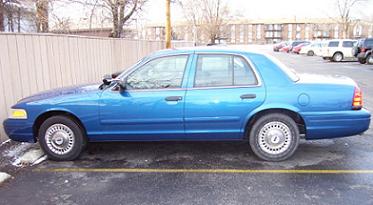 2000 Ford Crown Victoria - A blue 2000 Ford Crown Victoria sedan is parked in a lot with other vehicles visible in the background.