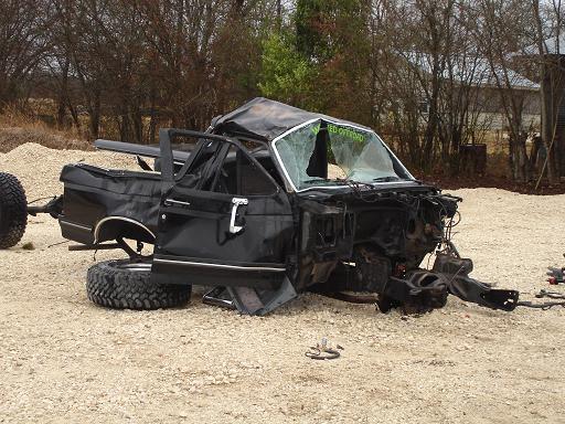 Wrecked black truck, front end missing, doors open, cracked windshield with text