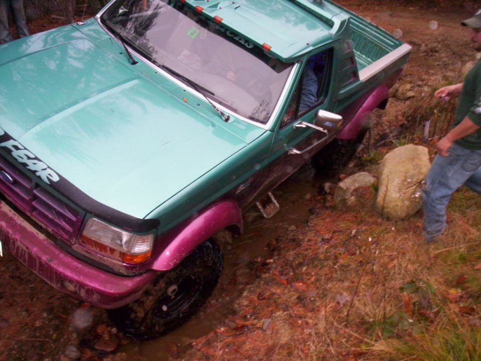 Teal and pink lifted Ford truck flexed in mud with a person standing beside it