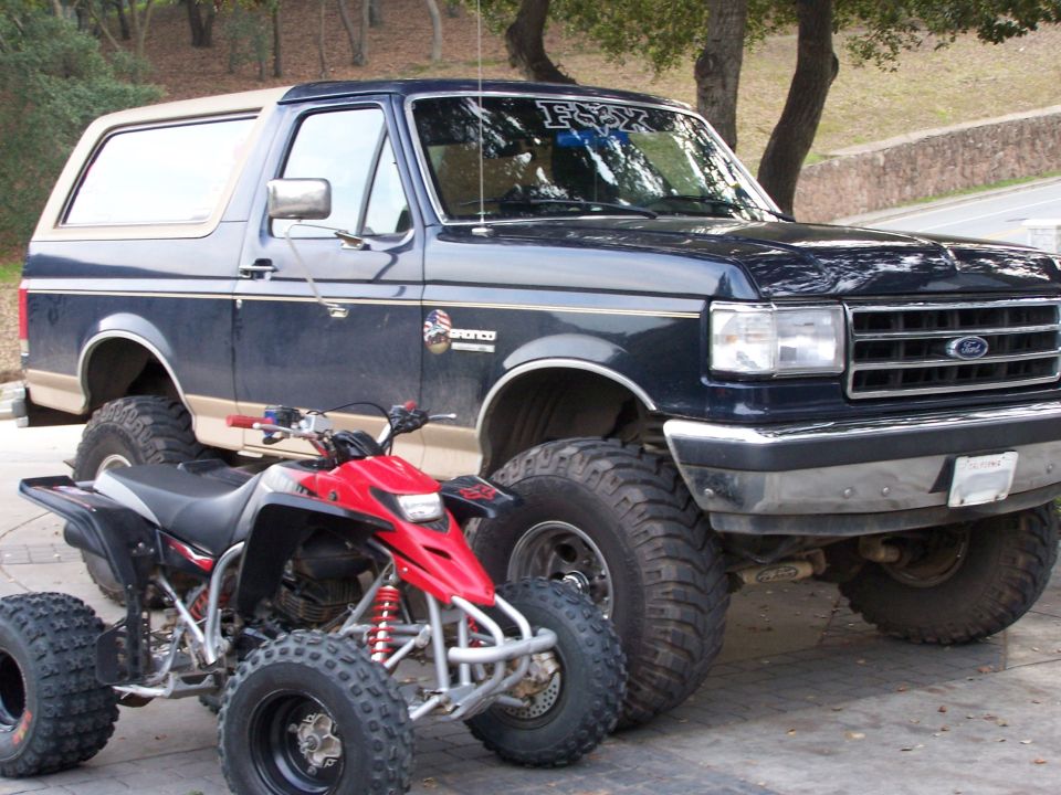 Lifted blue and tan Ford Bronco on mud tires with a red sport quad parked in front