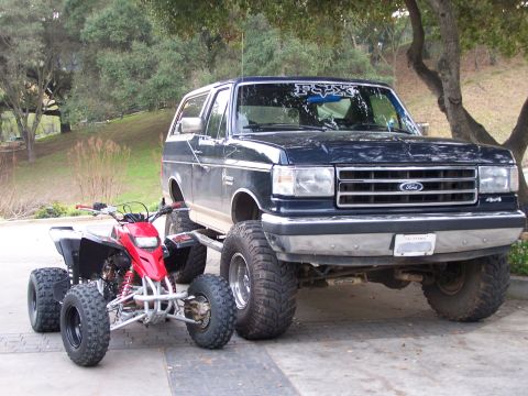 1991 Ford Bronco - A lifted 1991 Ford Bronco sits beside a red ATV on a paved surface with trees in the background.