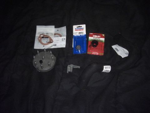 1991 Ford LTD Crown Victoria - A flat-lay of new tune-up and service parts on a dark blanket: a grey distributor cap with terminal towers, a bagged gasket set, a Purolato…