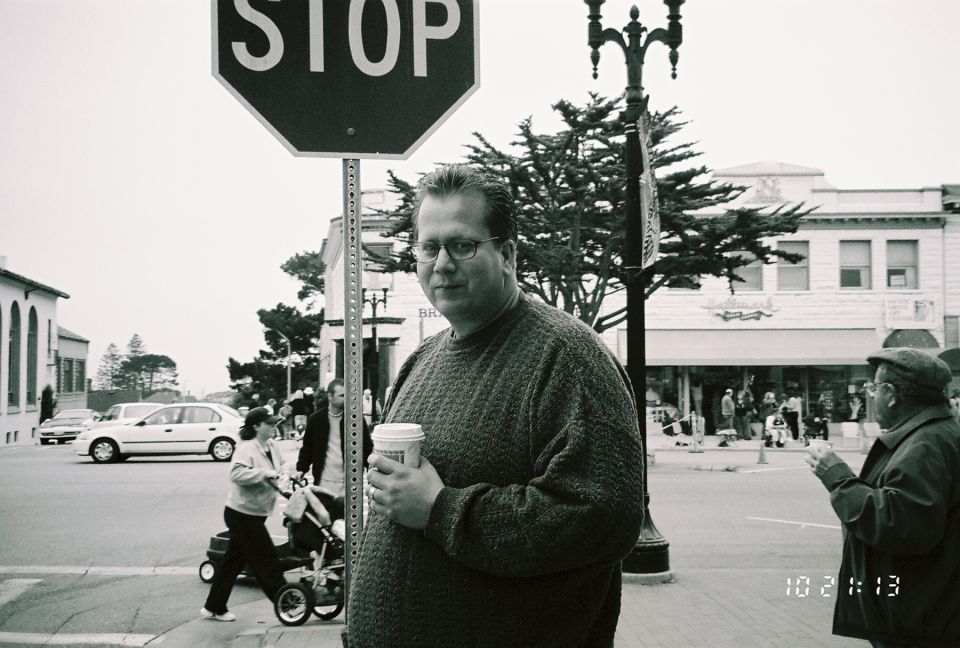 Man in sweater holding coffee cup in front of stop sign, with cars and buildings in background