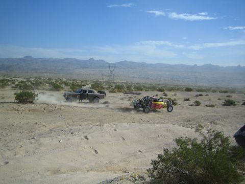 Dark sedan and red-yellow buggy racing across desert flats near power lines