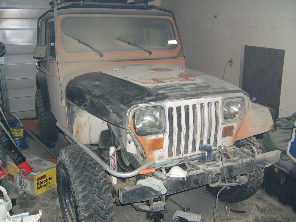 Jeep Wrangler YJ in garage mid-restoration with sanded hood, removed bumper, and tools nearby