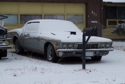 1972 Ford Ranchero - A weathered car sits covered in snow, its hood scoop visible, parked in front of a garage with a mailbox in the foreground.