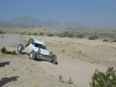 White open-frame off-road buggy catching air over a sand berm