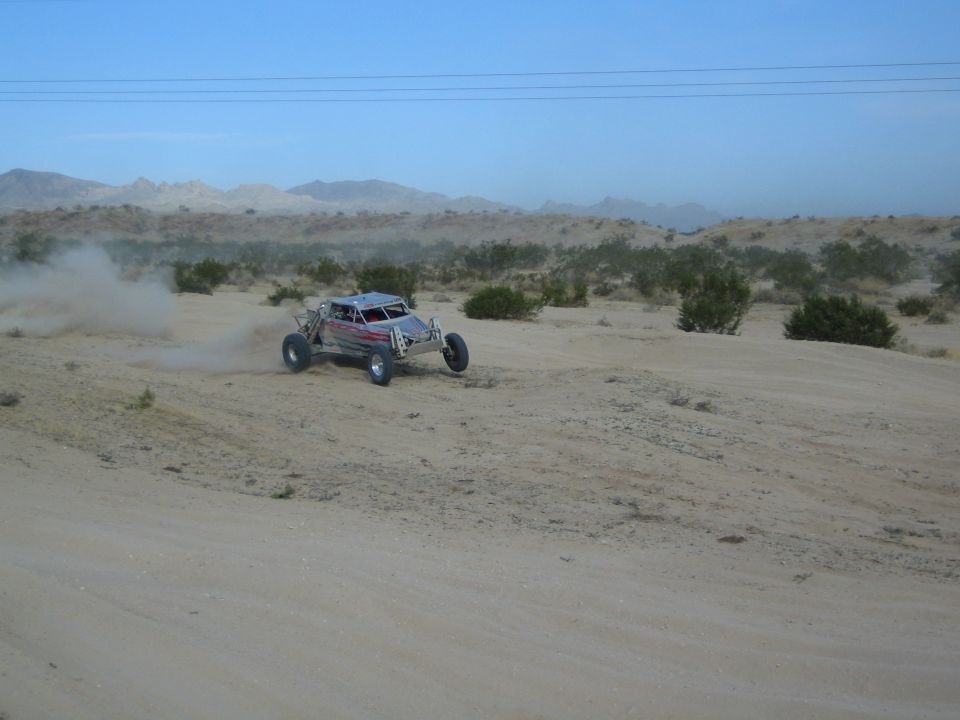 Silver off-road buggy racing across sandy desert with mountains behind