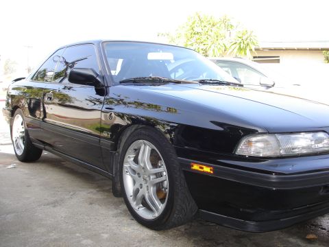 1989 Mazda MX-6 - A black 1989 Mazda MX-6 GT coupe is shown from the front 3/4 angle, highlighting its sporty design and alloy wheels.