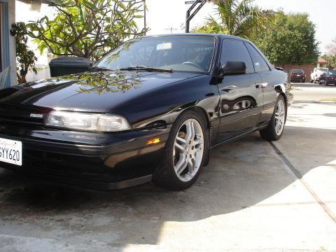 1989 Mazda MX-6 - A black 1989 Mazda MX-6 GT is parked on a driveway with trees and a house in the background.