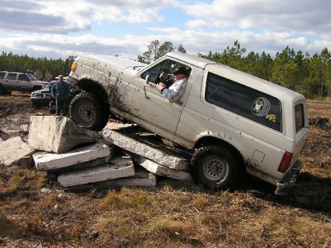 1989 Ford Bronco - White 1989 Ford Bronco is shown off-roading over concrete blocks, with another SUV in the background.