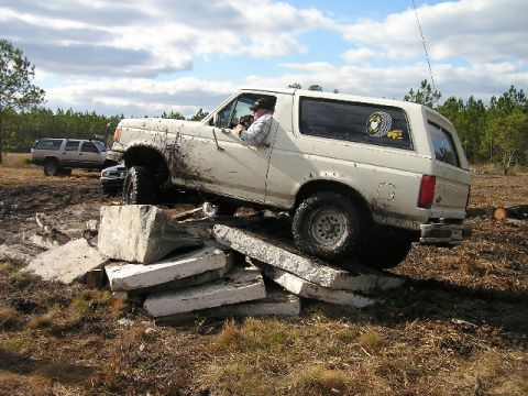 1989 Ford Bronco - Mud-splattered tan full-size Ford Bronco SUV with removable hardtop climbing a pile of broken concrete slabs at an outdoor off-road play ar…