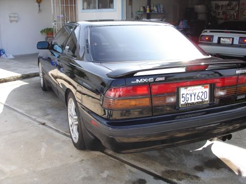 1989 Mazda MX-6 - This 1989 Mazda MX-6 4WS coupe is shown from the rear, parked in a driveway with another vehicle visible in the background.