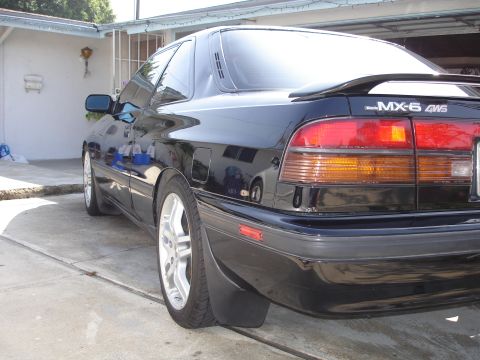 1989 Mazda MX-6 - The rear of a black Mazda MX-6 GT AWD is shown in a driveway, reflecting the surroundings.