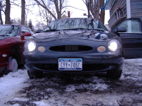 1998 Ford Taurus - A dark sedan is parked in a snowy area with its headlights illuminated. Another vehicle is partially visible to the left.