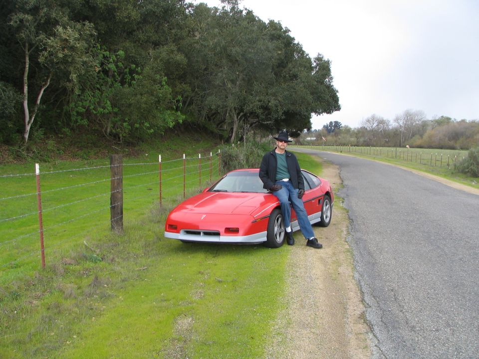 Man in cowboy hat leans on red 1986 Pontiac Fiero parked on roadside
