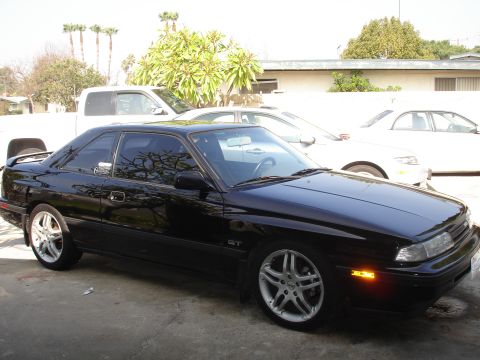 1989 Mazda MX-6 - A black 1989 Mazda MX-6 GT is parked in front of a building with other vehicles visible in the background.
