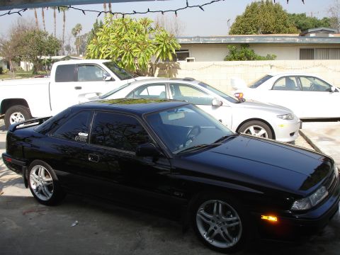 1989 Mazda MX-6 - A black 1989 Mazda MX-6 GT coupe is parked in front of other vehicles under a string of lights.