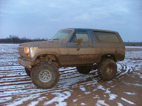 1985 Ford Bronco - This off-road vehicle is covered in mud and parked in a field with patchy snow under a cloudy sky.