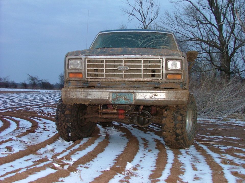 Mud-covered 1980s Ford Bronco front view, lifted on large tires, in a snowy field