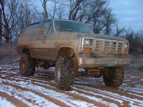 1985 Ford Bronco - This lifted SUV, likely from the early 1980s, is covered in mud and driving through a snowy, rutted field.