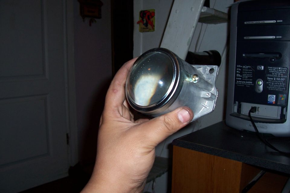 Headlight projector held in hand, chrome bezel, clear lens with burn marks, next to a computer tower