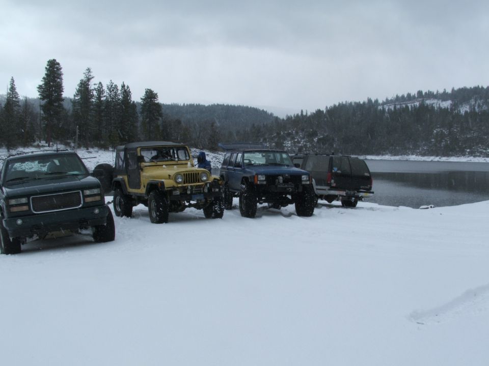 Yellow lifted Jeep CJ7 with black soft top parked in snow beside other 4x4s near a lake