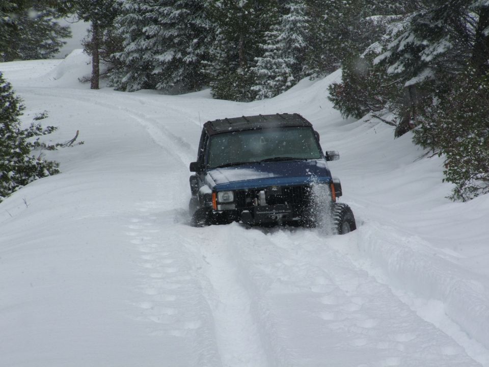 Dark blue Jeep SUV plowing through deep snow on a forested trail, snow caked on its front end