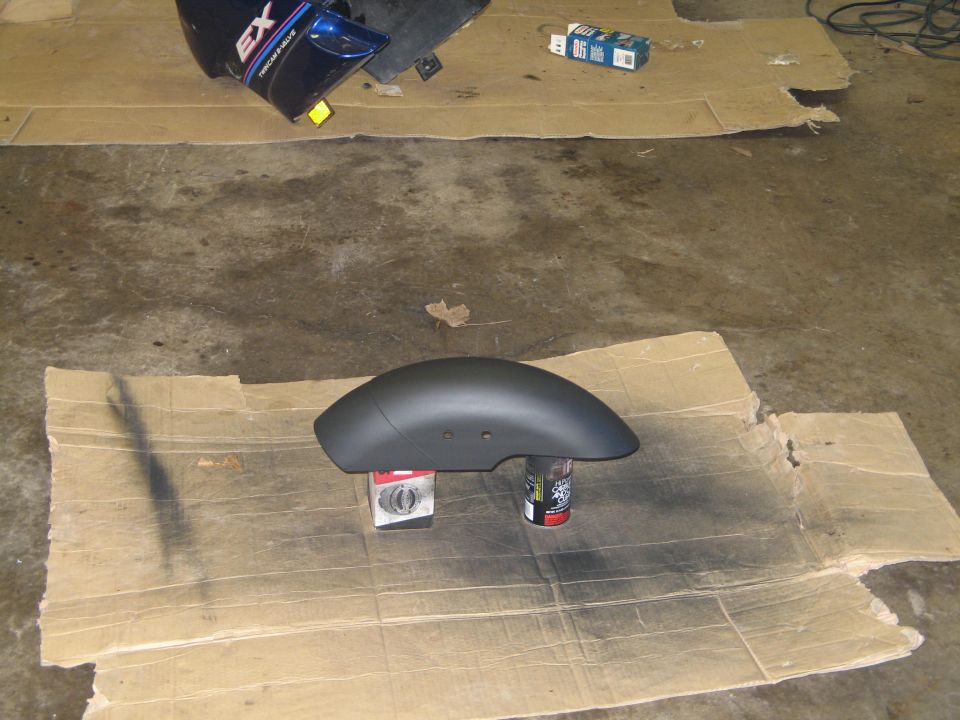 Motorcycle front fender painted flat black, drying on spray cans over cardboard in a garage