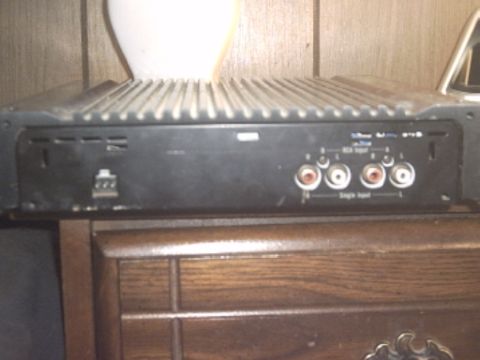 1991 Ford LTD Crown Victoria - A black finned-heatsink car audio amplifier sitting on a dark wood dresser indoors, photographed from the input end. Four RCA jacks labeled…