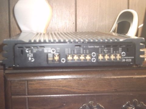 1991 Ford LTD Crown Victoria - A black finned car audio amplifier resting on a dark wood dresser, photographed along its output end where gold speaker-output and power te…