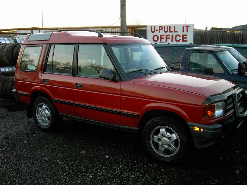 Red Land Rover Discovery SUV parked in a U-Pull-It salvage yard, front three-quarter view