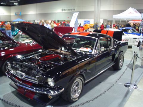 1984 Ford Bronco - A black 1965 Ford Mustang with racing stripes is displayed with its hood and trunk open at an indoor event.
