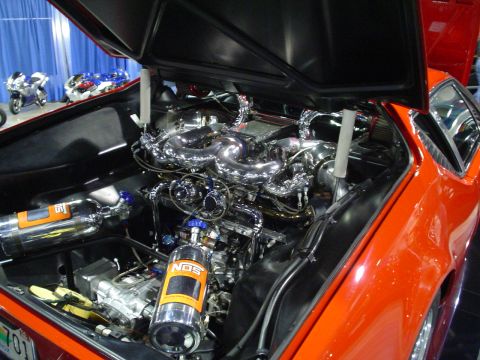 1984 Ford Bronco - Engine bay of an orange car with chrome components and NOS tanks is visible, with motorcycles in the background.