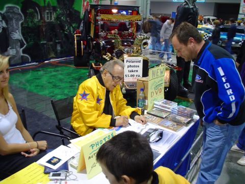 1984 Ford Bronco - People interact at a booth during a car show event, with merchandise displayed on the table.