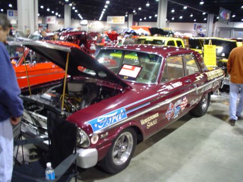 1984 Ford Bronco - A maroon 1960s Ford sedan, likely a Falcon, is displayed with its hood open at a car show, racing-themed decals.