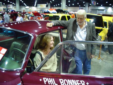 1984 Ford Bronco - A man and woman stand by a maroon car at a car show with other vehicles in the background.