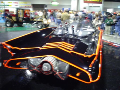 1984 Ford Bronco - A custom car with a unique design and orange accents is displayed indoors with other vehicles and people.