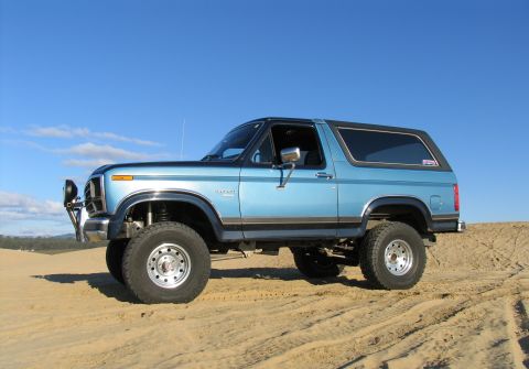 1984 Ford Bronco - This lifted 1984 Ford Bronco is shown in a 3/4 front view on a sandy terrain under a clear blue sky.