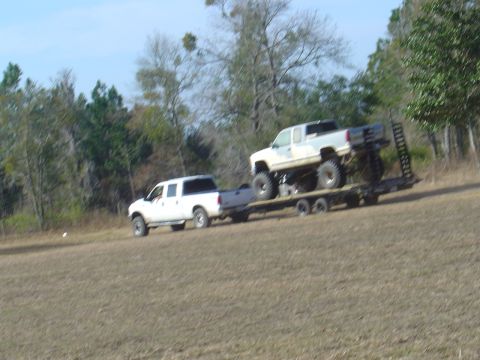 1987 Suzuki Samurai - A white pickup truck tows a trailer carrying a heavily modified lifted truck through a grassy field.