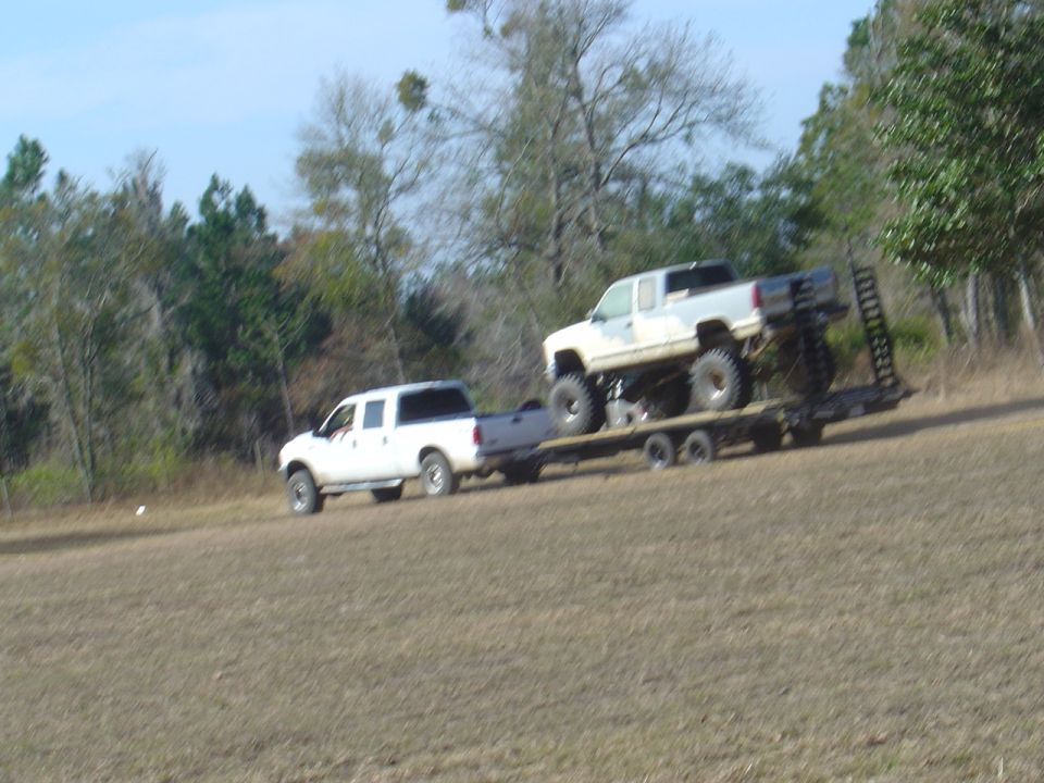 White pickup truck towing a trailer with a lifted truck on it